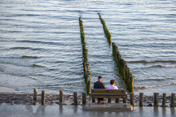 An elderly couple sitting on a bench on the beach, at high tide, breakwater, Netherlands