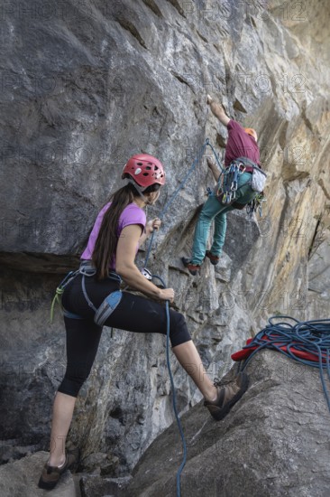 A woman stands at the base of a mountain, holding a climbing rope as her partner ascends to the top