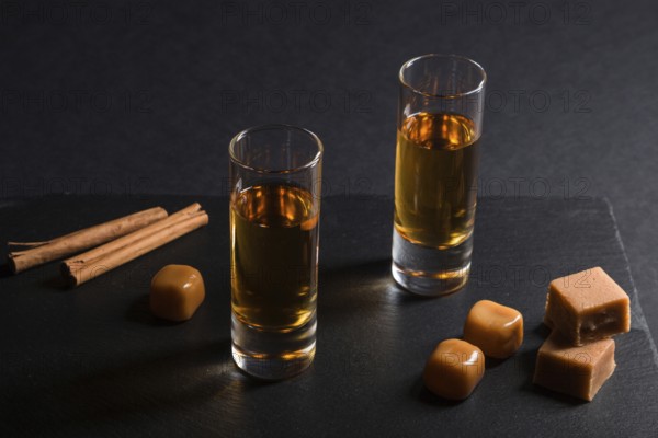 Glass of whiskey with caramel candies on a black stone slate board on black background. Side view,