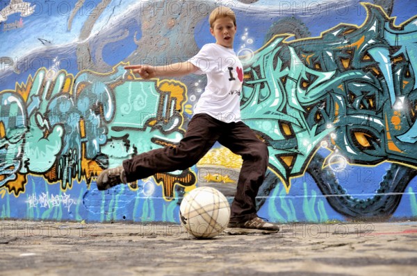 A ten-year-old boy plays with his football in front of a graffiti wall, football pitch in Germany