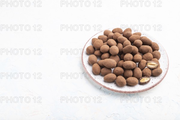 Almond in chocolate dragees on ceramic plate on white concrete background. Side view, copy space,