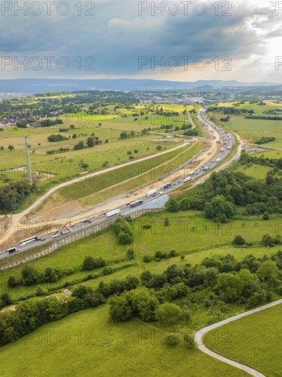 View of a large construction site next to a road through green fields and under a cloudy sky, A8