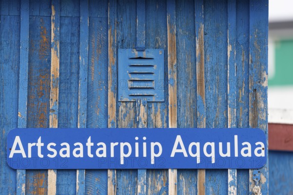 A blue sign inscribed with a Greenlandic street name on a weather-beaten wooden wall, remote Arctic