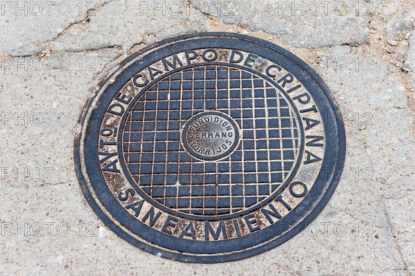 A round metal manhole cover with text engraving on a tarmac road, manhole cover, Campo de Criptana,