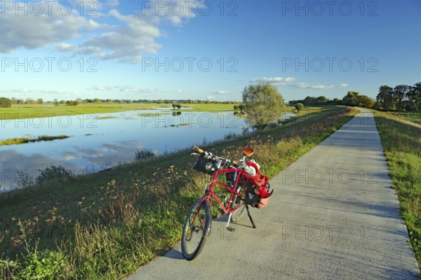 A bicycle stands on a concrete path next to a calm river under a clear blue sky with scattered