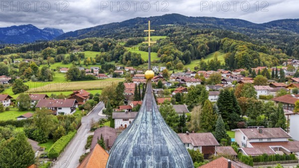 Village view with church spire cross, surrounded by green hills and wooded mountains, under a