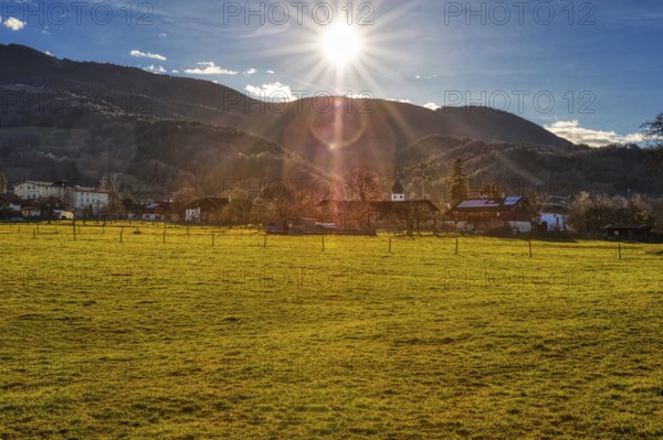 The sun sets over a village that lies in front of a mountain range and is surrounded by green