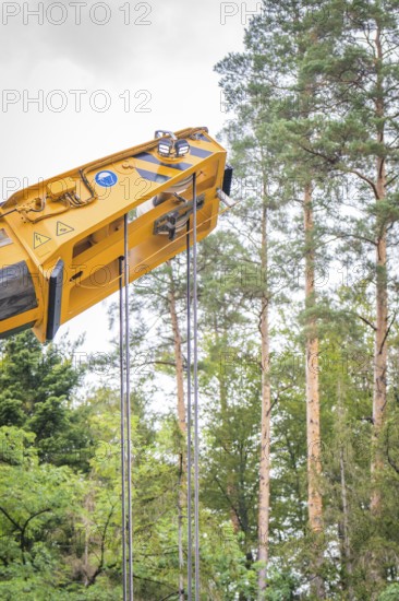 Yellow crane arm with ropes in front of a background of tall trees and sky, track construction,
