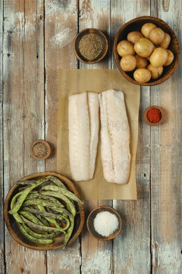 Two fresh cod fish fillets on parchment paper surrounded by various spices, green beans, and small