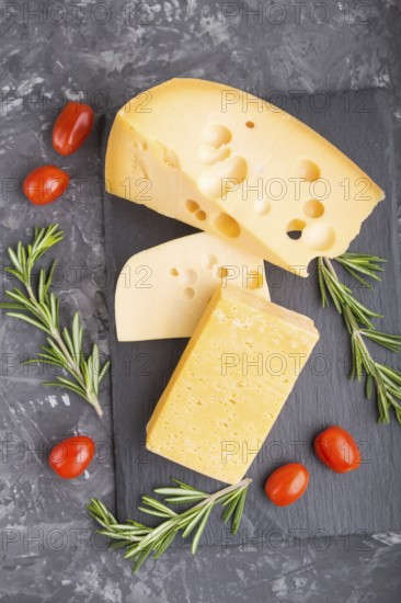 Various types of cheese with rosemary and tomatoes on black slate board on a black concrete