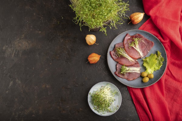 Slices of smoked salted meat with cilantro microgreen on black concrete background and red textile.