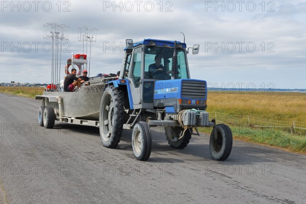 A blue tractor on a country road with people in the trailer, rural surroundings, tractor with boat