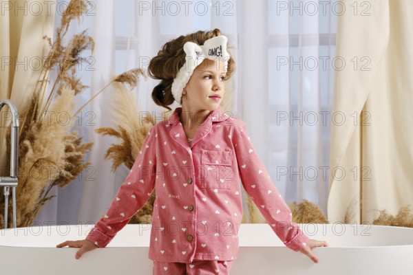 Cute child in a pink pajama and hair band stands next to a modern white bathtub spreading hands