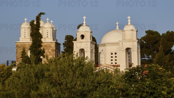 White church with three domes and bell towers, surrounded by greenery under a clear sky, Koroni,
