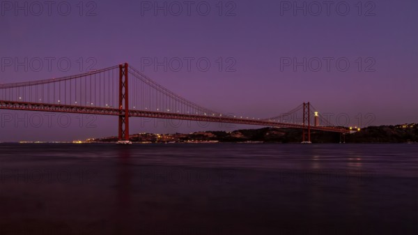 Twilight descends on a calm river with a suspension bridge standing majestically against a purple