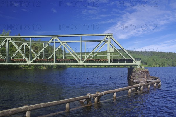 A green metal bridge crosses a river in a natural setting with trees and blue sky, railway bridge,