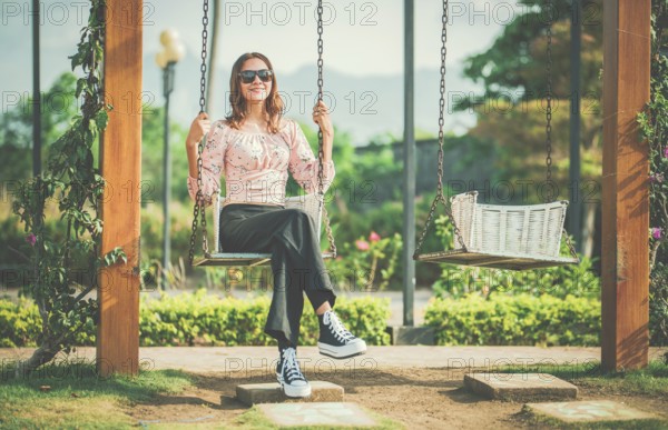 Cheerful girl in glasses relaxing on a swing in a natural park. Happy young woman sitting on a