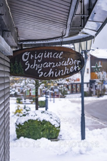 Company sign for Black Forest ham on a snowy street with shops, Enzklösterle, district of Calw,
