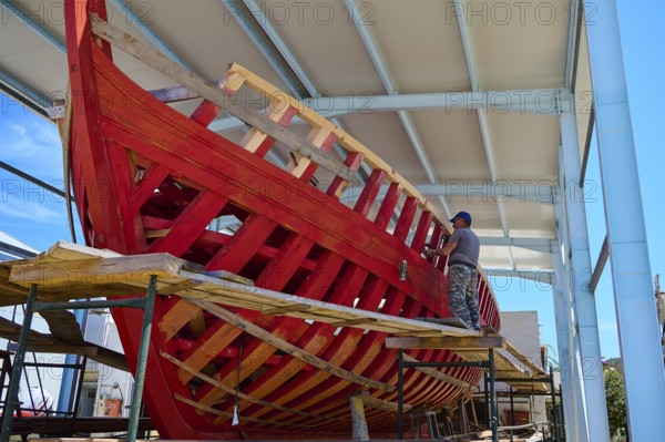 A partially built red wooden ship in a covered shipyard with a labourer working on it, Patmos