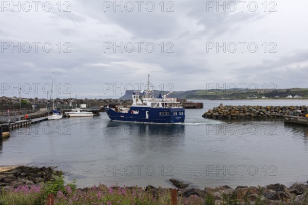 A blue ship sails in a cloudy harbour with rocky shores and blue water, Ballycastle