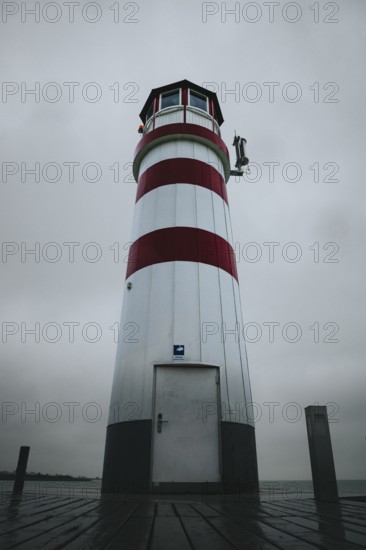 A red and white lighthouse stands on a wooden plank under a grey sky, Podersdorf, Burgenland,