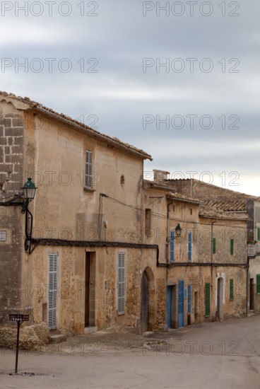 Medieval village street with stone buildings and colourful shutters under a cloudy sky, Sineu,