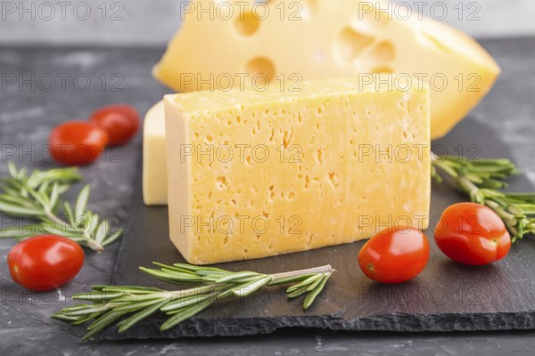 Various types of cheese with rosemary and tomatoes on black slate board on a black concrete