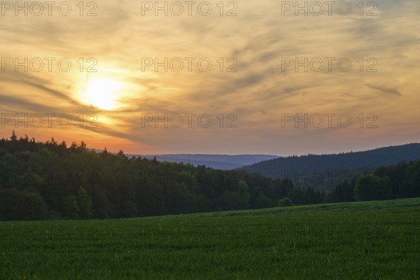 Sunset over a green meadow, surrounded by forest and hills under a cloudy sky, summer, Mönchberg,
