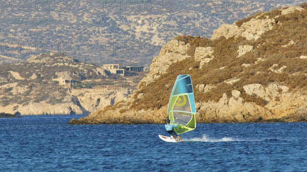 Person windsurfing along a rocky coast on blue water, windsurfer, Meltemi windsurfing spot, Devils