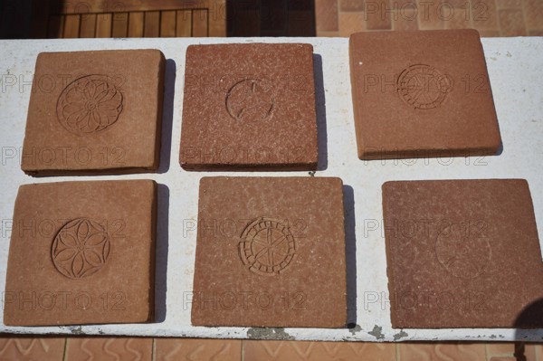 Six square clay bricks with different geometric and floral patterns laid on a white table top,