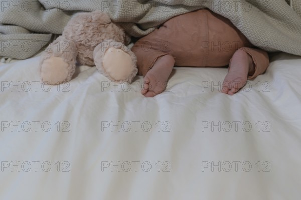 Little baby in brown trousers, next to a plush toy under a blanket on a bed