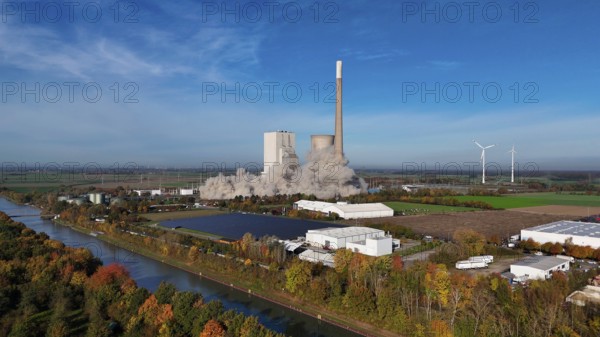 Demolition of a chimney with smoke cloud, surrounded by autumn landscape and wind turbines,