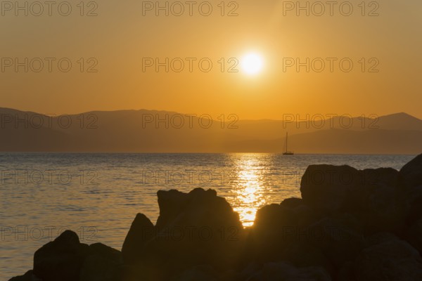 Sunset over the sea with a sailing boat in the distance, rocks in the foreground, Agíos Nikólaos,