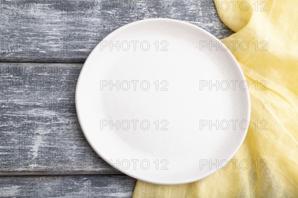 Empty white ceramic plate on gray wooden background and yellow linen textile. Top view, close up,