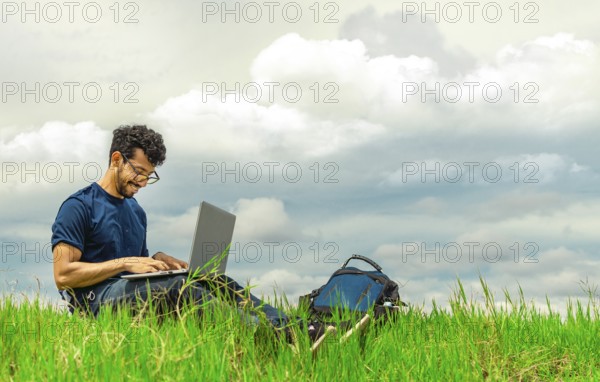 Smiling freelancer man sitting with laptop and backpack in field. Portrait of a man sitting on the