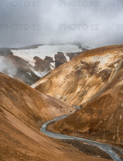 Steaming stream between colourful rhyolite mountains with snowfields, Hveradalir geothermal area,
