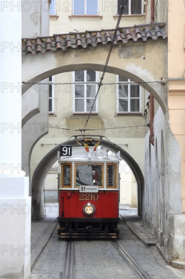 Red historic tram runs through winding houses and alleys with archways in Prague, Czech Republic,