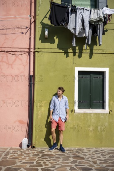 Young man leaning against a house wall, green and pink house facade with laundry to dry, colourful