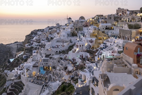 Cliff-side houses, villas and windmill in the village of Ia seen from Oia Castle at dusk,
