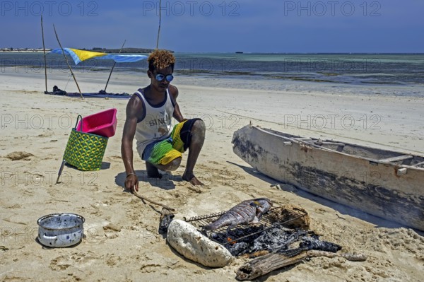 Malagasy man cooking freshly caught fish on grill over charcoal fire on sandy beach at Andavadoaka