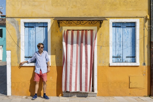 Young man leaning against house wall, yellow house facade with entrance door with red and white