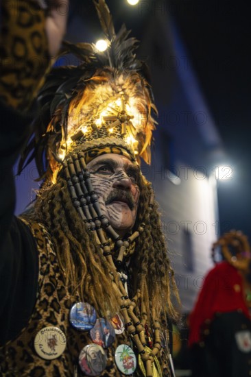 Man in festive, feathery costume with illuminated hat and decorated buttons at night event,