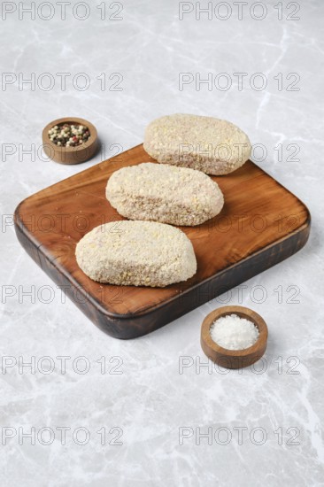 Three breaded cutlets on a rustic wooden cutting board, accompanied by small bowls of salt and