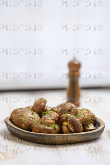 A wooden plate filled with roasted mushrooms garnished with herbs on a rustic table. A pepper mill