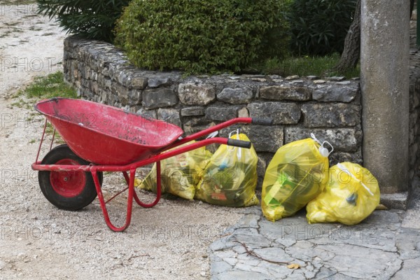Red metal wheelbarrow next to translucent yellow plastic thrash bags filled with various discarded