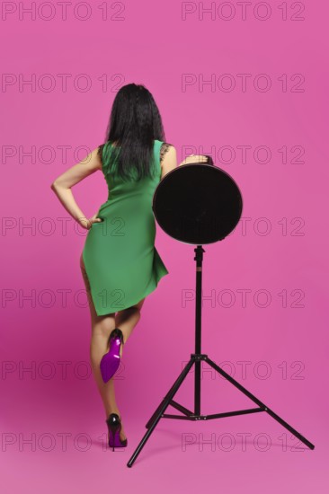 Rear view of woman with black hair and in green dress rising one leg and leaning on studio strobe