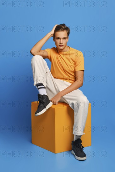 Positive man in orange undershirt and white sweatpants sits on wooden cube over blue studio