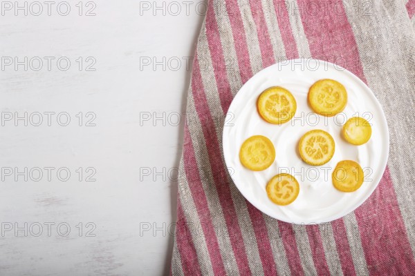 Greek yogurt with kumquat pieces in a white plate on a white wooden background, top view, flat lay,
