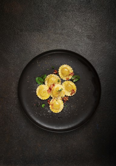Fresh ravioli, with butter, spices and herbs, fried bacon, on a black plate, top view, dark