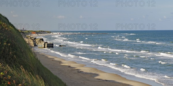 View of the beach with gentle waves lapping against the shore, overgrown with grass and sand dunes,